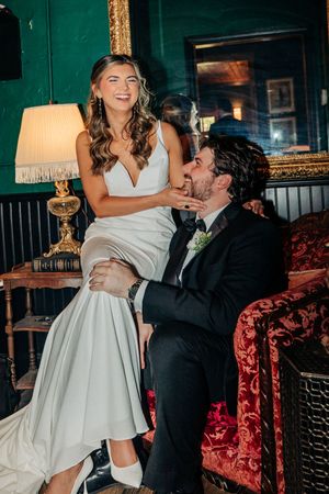 Newlywed couple shares joyful moment on vintage red velvet couch with bride in white dress and groom in black suit.