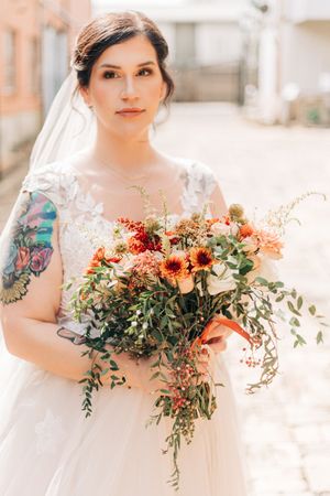 A bride in a white lace wedding dress holds a vibrant autumn bouquet with sunflowers and trailing greenery.