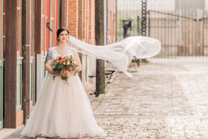 A bride stands on a brick sidewalk in a flowing white gown with a long veil caught gracefully in the wind.