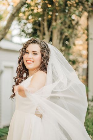 A bride in a white wedding dress with flowing curly hair and veil poses outdoors during an autumn photoshoot.