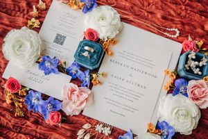 Wedding invitation suite displayed with colorful fresh flowers in blue, coral, and white on a red textured background.