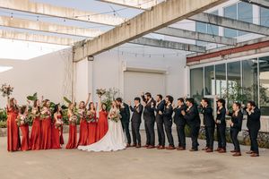 Wedding party lined up outside modern building with bridesmaids in red dresses and groomsmen in dark suits.