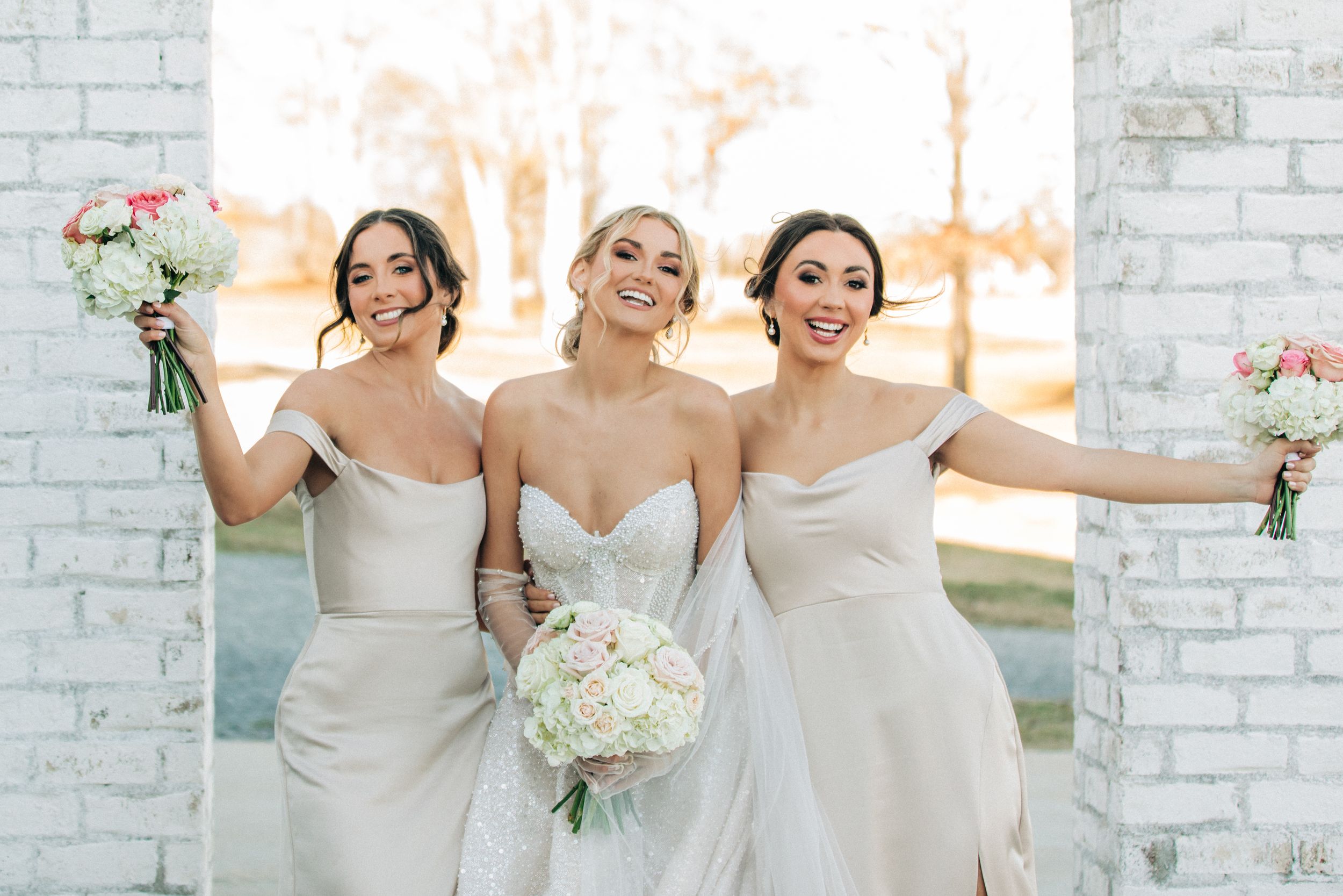 Three bridesmaids in elegant white dresses pose joyfully while holding floral bouquets against a white brick wall.