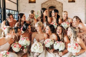 A wedding party wearing blush bridesmaid dresses and holding floral bouquets share laughter together against a brick wall.