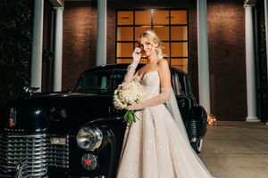 Bride in an elegant strapless ballgown poses at twilight against a grand brick building with illuminated windows.