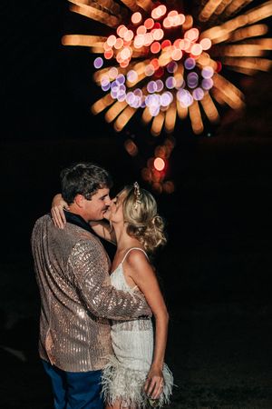 A couple shares a romantic kiss under glowing pink bokeh lights at a nighttime wedding celebration.
