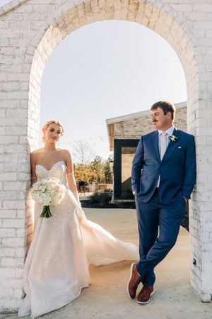 A newlywed couple poses under a white brick archway, with the bride in a strapless gown and groom in navy suit.