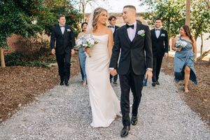 Wedding party walks along gravel path with couple in white dress and black tuxedo leading the group at outdoor venue.