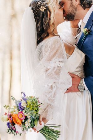 A romantic wedding couple sharing a kiss while the bride holds a colorful summer bouquet of wildflowers.