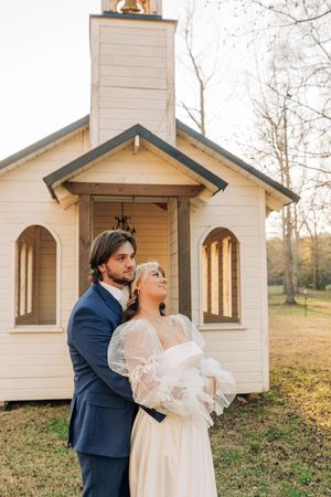 A couple shares an intimate moment outside a charming white wooden chapel on an autumn day.