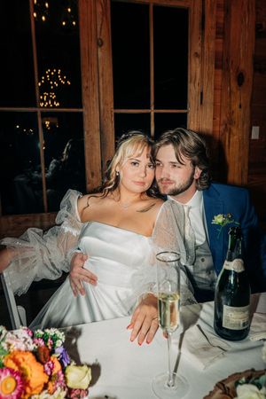 Newlywed couple shares intimate moment at rustic reception with champagne and colorful floral centerpiece.