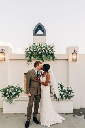 Newlywed couple sharing an intimate moment outside a white chapel decorated with elegant floral arrangements and lanterns.