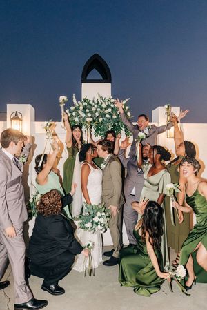 Wedding guests dance enthusiastically in a line during reception celebration with green decor and white floral arrangements.