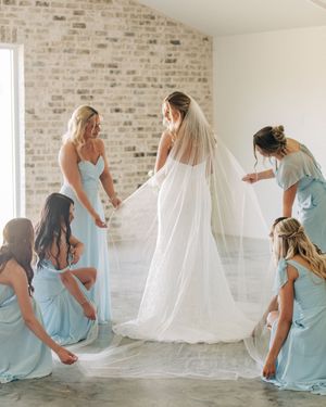 Bridesmaids in light blue dresses help adjust the bride's long flowing veil against a rustic brick wall background.