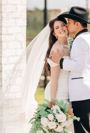Newlyweds share a tender moment outdoors as groom kisses bride holding white and green floral bouquet.