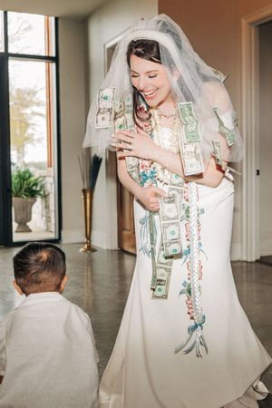 A bride in a white wedding dress laughs while holding dollar bills attached to her gown in a money dance tradition.
