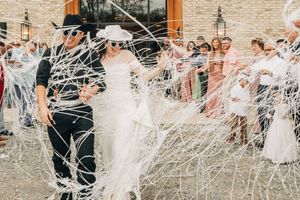 Wedding guests throw white stringy confetti over a couple exiting their ceremony on a sunny day outside a brick building.