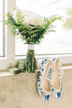 White high heel wedding shoes with blue floral designs displayed in a bright window setting with flower bouquets.