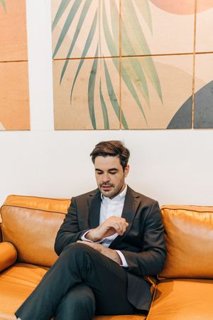 Business professional in dark suit checks device while seated on tan leather couch in modern office lobby.