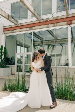A couple embraces in front of a modern glass building with tall ornamental grasses in a wedding photo.