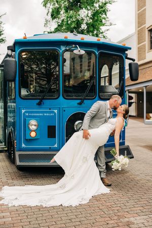 A couple shares a romantic dip kiss in front of a bright blue vintage trolley on a brick paved street.