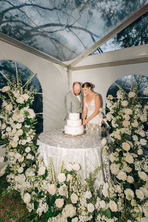 A couple stands at their wedding cake table under a white tent surrounded by elegant white floral arrangements.