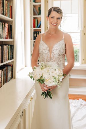 A stunning bride in a plunging V-neck lace gown holds a white bouquet while standing in a library setting.