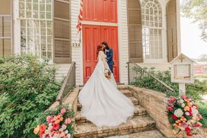 Couple stands by bright red church door surrounded by floral arrangements and greenery in romantic wedding scene.
