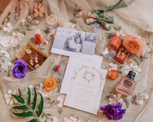 Flatlay of wedding stationery with purple and coral flowers, perfume bottle, and greenery on textured fabric background.