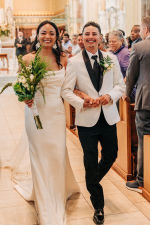 Happy newlyweds walk down the aisle of an ornate church, bride in elegant white dress holding bouquet, groom in white jacket.