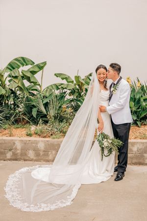 A couple shares a romantic moment on their wedding day in front of lush tropical greenery and a flowing cathedral veil.