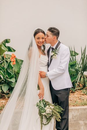 Newlyweds share intimate moments in a garden setting with tropical plants and flowing white veil.
