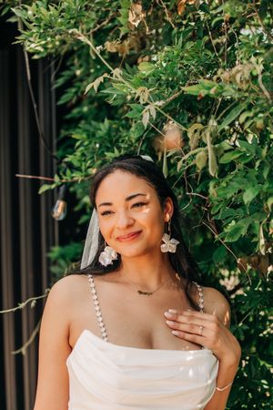 A radiant bride wearing a white strappy dress poses against lush green foliage in an outdoor portrait session.