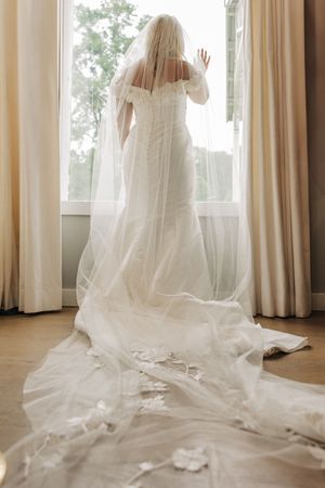 Back view of elegant bridal gown with flowing cathedral veil against sunlit window in minimalist room.