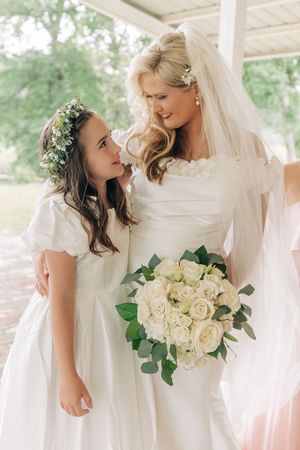 A bride in white dress holds a cream rose bouquet with eucalyptus while sharing a moment with flower girl in matching white.