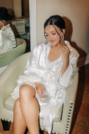 Woman in luxurious white silk robe and nightwear sitting elegantly on upholstered chair while touching earring.