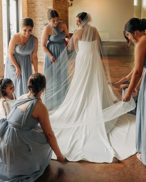 Bridesmaids in dusty blue dresses help arrange the bride's flowing cathedral veil in a rustic venue.