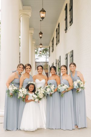 Wedding party in elegant light blue bridesmaids dresses posing on elegant white columned porch with hanging lanterns.