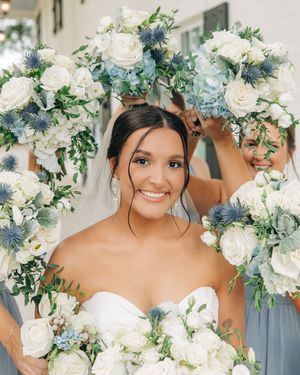 Beautiful wedding bouquet featuring white roses and blue thistles with delicate greenery held by a joyful bride.