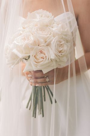 A delicate bouquet of cream-colored roses wrapped in white stems held through soft flowing bridal veil fabric.