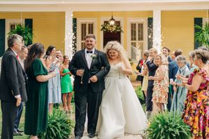 A joyful wedding party exits while guests celebrate on the porch of a yellow house decorated with white columns and greenery.