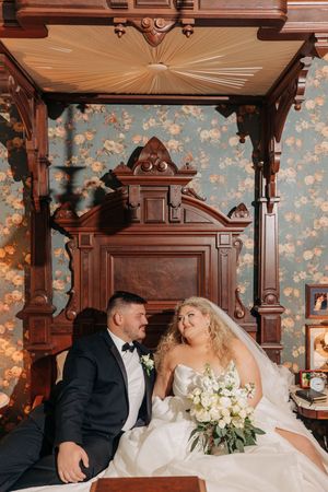 A couple sits in an ornate antique wooden chair against floral wallpaper with white bouquet and wedding attire.