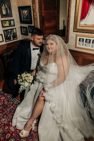 Elegant wedding couple sitting on an ornate carpet surrounded by framed photos on wooden walls during intimate ceremony.