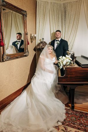 Beautiful wedding scene with newlyweds posing by antique piano and ornate mirror in elegant Victorian-style room.