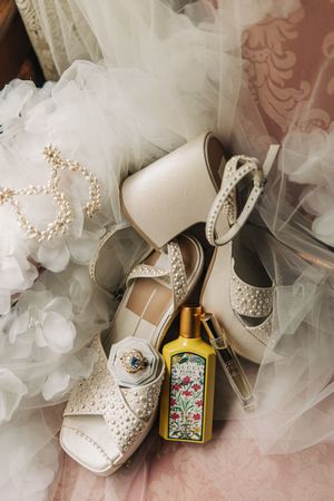 Wedding accessories including white lace shoes, pearl jewelry, tulle veil and decorative perfume bottle arranged on pink fabric.