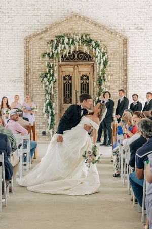 A panoramic view of a white brick chapel wedding ceremony showing guests seated and an ornate floral arch at the altar.