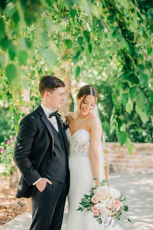 A couple in wedding attire poses together under hanging greenery in a romantic garden setting with soft natural light.