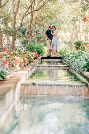 Couple standing on ornate garden water feature with cascading fountains surrounded by lush flowers and trees.