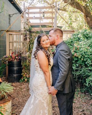 A loving wedding couple embraces in a romantic garden setting with natural greenery and wooden structures.