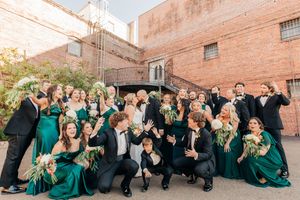 A wedding party wearing emerald green dresses and black suits poses together outdoors against a brick wall.